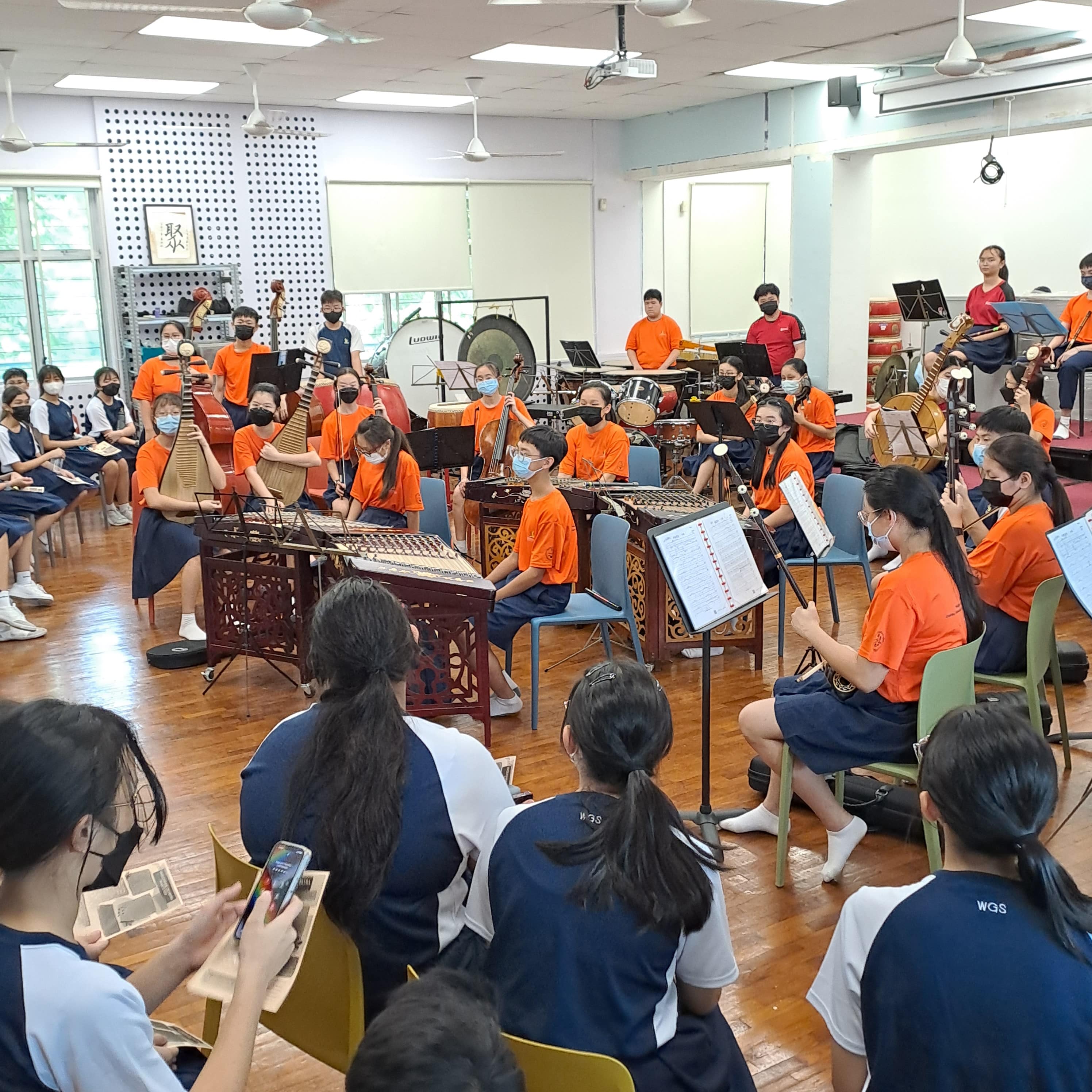 Students in orange shirts play Chinese instruments in a classroom while others in blue uniforms watch.